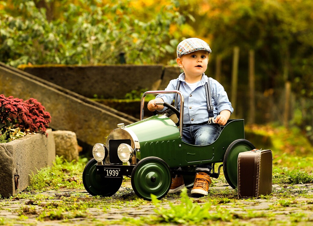 3jähriger Junge in einem grünen Garten in einem dunkelgrünen Spielzeugauto und braunen Spielzeugkoffer